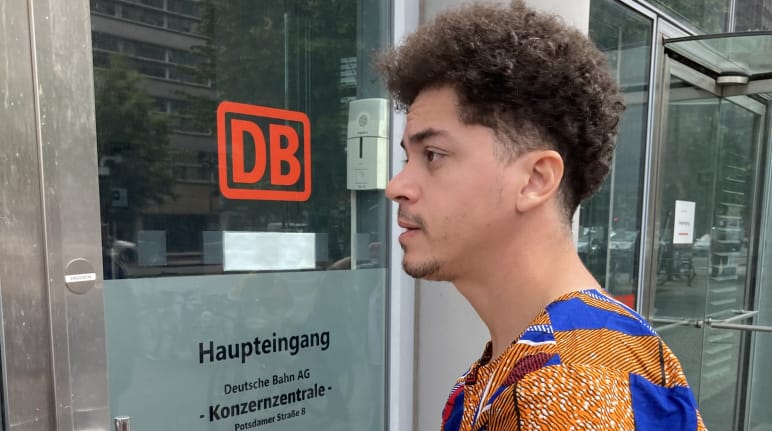 Un jeune homme se tient avec une enveloppe devant une porte vitrée portant l’inscription DB entrée principale siège du groupe Deutsche Bahn AG
